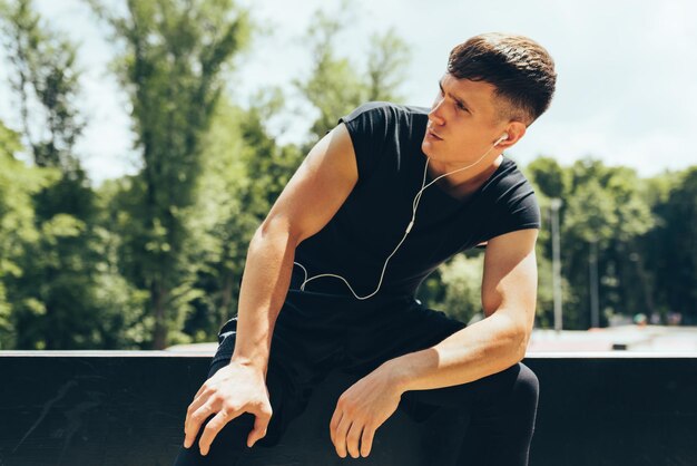 Horizontales Porträt eines jungen Mannes, der sich nach dem Training ausruht, um die Musik über Kopfhörer im Freien zu hören Junge muskulöse Männer ruhen sich nach einem harten Training auf dem Sportplatz aus Menschen- und Sportkonzept