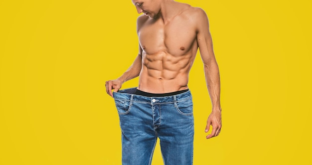 Horizontales Studiobild des jungen kaukasischen fitten Mannes, der nach der Diät große blaue Jeans trägt und sexy Torso zeigt Attraktiver Mann, der sich nach dem Abnehmen auf gelbem Hintergrund ausgibt Kopieren Sie Platz Gesunder Lebensstil