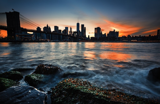 Horizonte de Manhattan com a ponte de Brooklyn à noite. Rochas e pedras na margem do East River