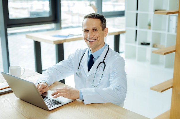 Hospital moderno. belo e simpático médico inteligente sentado à mesa e sorrindo enquanto usa seu laptop