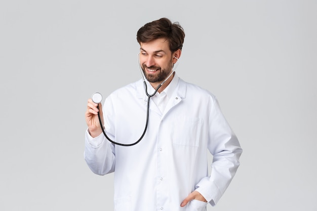 Hospital, profissionais de saúde, conceito de tratamento covid-19. Médico bonito com barba em uniforme branco, usando estetoscópio para ouvir os pulmões do paciente, tratar clientes na clínica, sorrindo, plano de fundo cinza