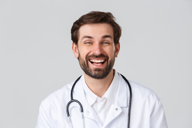 Hospital, trabajadores sanitarios, concepto de tratamiento covid-19. Médico barbudo alegre con actitud positiva, tomando un descanso, sonriendo y riendo mientras habla, usa matorrales, fondo gris de pie.