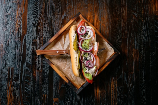 Hot Dog mit Zwiebel-Tomaten-Gurke und Sauce auf einem Holzständer, serviert mit einem Messer Fast Food mit Restaurant serviert