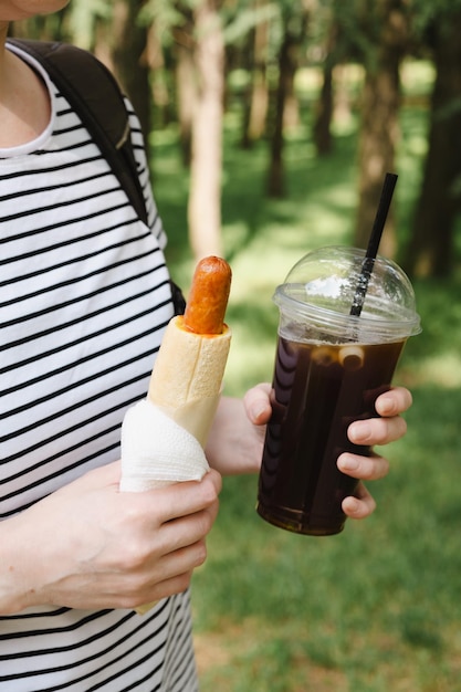 Hot Dog und schwarzer Eiskaffee in den Händen einer Frau Mittagessen während eines Sommerspaziergangs im Stadtpark