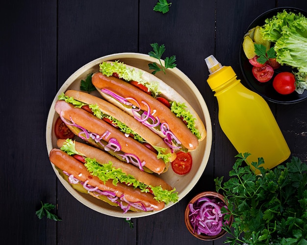 Hotdog mit Würstchen, Gurken, Tomaten, roten Zwiebeln und Salat auf dunklem Holzhintergrund