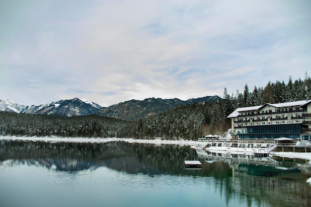 Hotel am See im europäischen Alpenwald im Winter mit Schnee und gefrorenen Bäumen und Bergen im Hintergrund