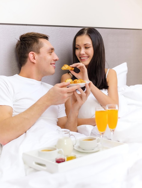 Foto hotel, viajes, relaciones y concepto de felicidad - pareja sonriente desayunando en la cama en la habitación del hotel