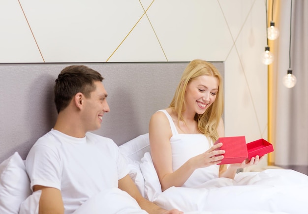 hotel, viajes, relaciones, vacaciones y concepto de felicidad - pareja sonriente en la cama con caja de regalo roja