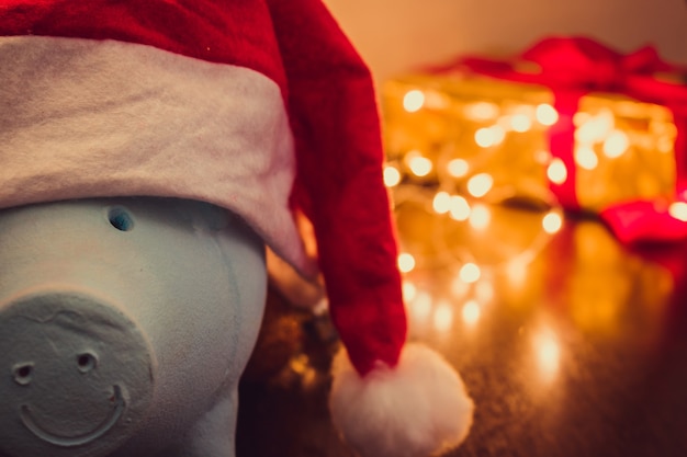 Hucha con sombrero de Santa Claus sobre fondo de Navidad borrosa con adornos navideños, regalos y luz. Espacio para texto.