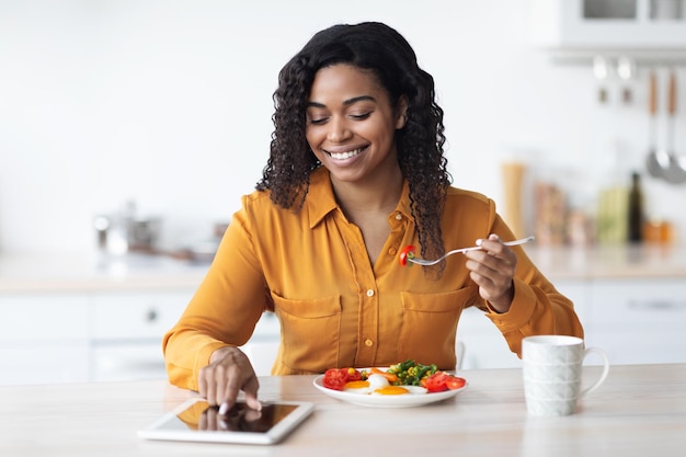 Hübsche afrikanisch-amerikanische Frau, die mit einem digitalen Tablet-Modell frühstückt