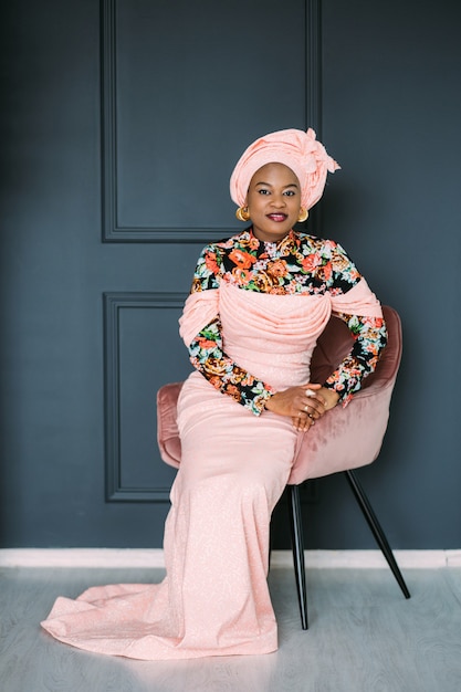 Hübsche afrikanische Frau, die ein rosa modisches traditionelles Kleid und ein Kopftuch trägt, auf dem Stuhl im Studio sitzt und mit einem Lächeln in die Kamera schaut. Dunkelblauer Wandhintergrund