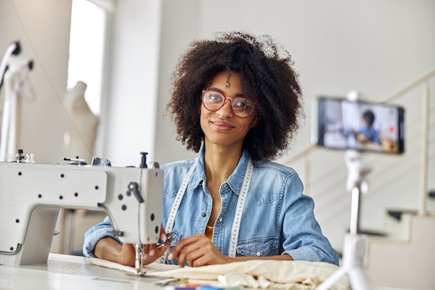 Hübsche afroamerikanische Bloggerin mit Schere und Brille am Arbeitsplatz in der Nähwerkstatt