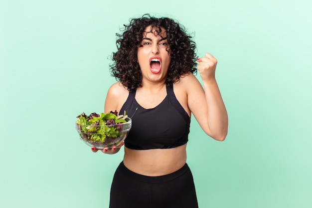Hübsche arabische Frau, die aggressiv mit einem wütenden Ausdruck schreit und einen Salat hält. Diätkonzept