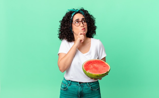 Hübsche arabische Frau, die denkt, sich zweifelhaft und verwirrt fühlt und eine Wassermelone hält. Sommerkonzept