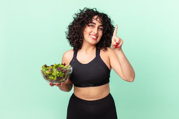 Hübsche arabische Frau, die freundlich lächelt und aussieht, Nummer eins zeigt und einen Salat hält. Diätkonzept