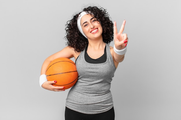 Hübsche arabische Frau, die glücklich lächelt und schaut, Sieg oder Frieden gestikuliert und einen Basketballball hält. Sportkonzept