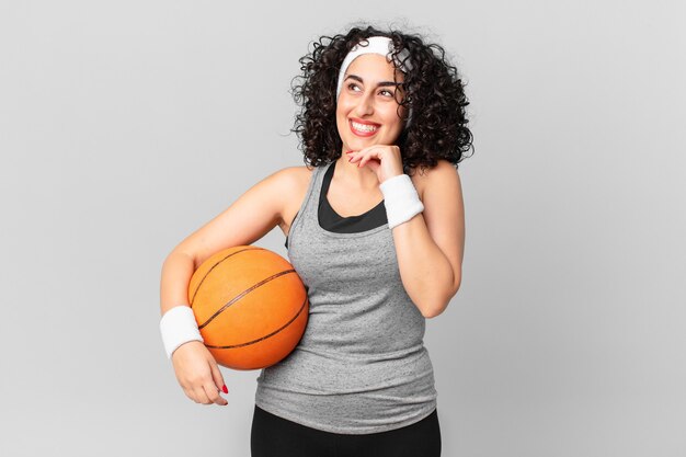 hübsche arabische frau, die mit einem glücklichen, selbstbewussten ausdruck mit der hand am kinn lächelt und einen basketballball hält. Sportkonzept