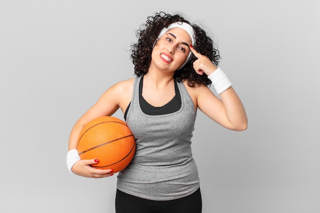 Hübsche arabische Frau, die sich verwirrt und verwirrt fühlt und zeigt, dass Sie verrückt sind und einen Basketballball halten. Sportkonzept