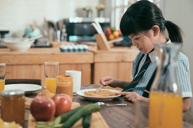 Hübsche asiatische chinesische kleine Studentin trinkt morgens gerne frischen Orangensaft beim Frühstück am Küchentisch zu Hause. Junges weibliches Kind, das im Speisesaal sitzt, legte Erdnussmarmelade auf Toast