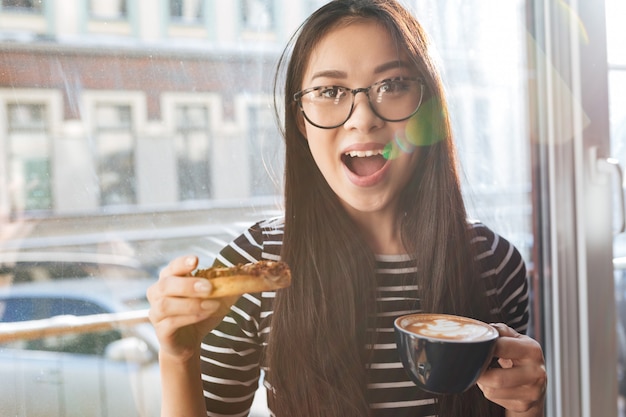 Hübsche asiatische Frau, die Kuchen auf Fensterbank isst