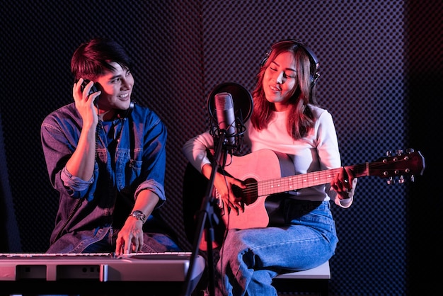 Hübsche asiatische Sängerin, die akustische Gitarre mit dem Mann spielt, der elektrische Tastatur spielt. Aufnehmen von Songs mit einem Studiomikrofon und Popschutz am Mikrofon in blauem und rotem Licht. Duett-Sitzung.
