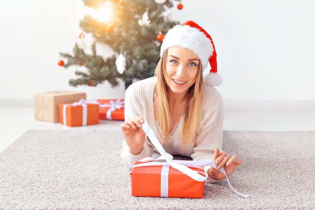 Hübsche blonde Frau, die einen warmen Strickpullover trägt und auf einem Teppich in der Nähe eines geschmückten Weihnachtsbaums in einem Wohnzimmer liegt. Viele Geschenke unter Baum.