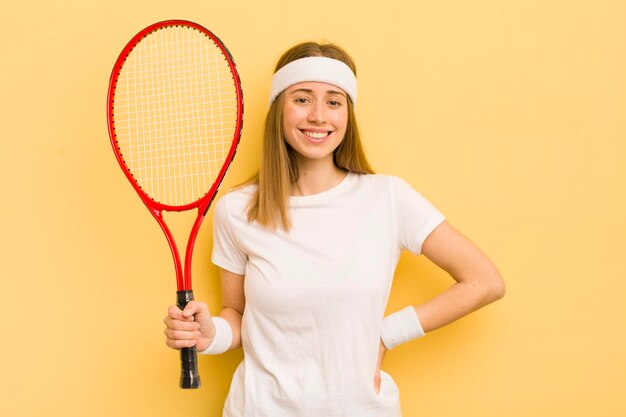 Hübsche blonde Frau, die glücklich mit einer Hand auf Hüfte und selbstbewusstem Tenniskonzept lächelt