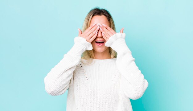 Hübsche blonde Frau, die lächelt und sich glücklich fühlt, die Augen mit beiden Händen bedeckt und auf eine unglaubliche Überraschung wartet