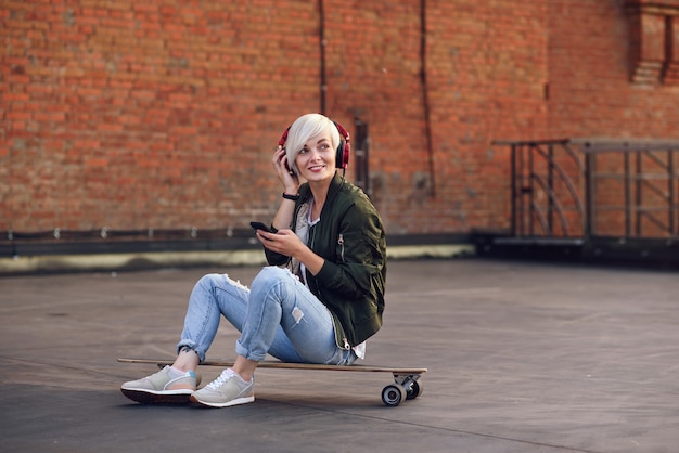 Hübsche blonde Frau, die Musik in den roten Kopfhörern hört und Telefon benutzt, während auf dem Longboard an der Backsteinmauer sitzt.