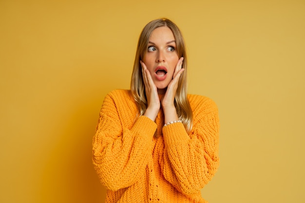 Hübsche blonde Frau mit überraschendem Gesicht im orange stilvollen Herbstpullover, der auf Gelb aufwirft.