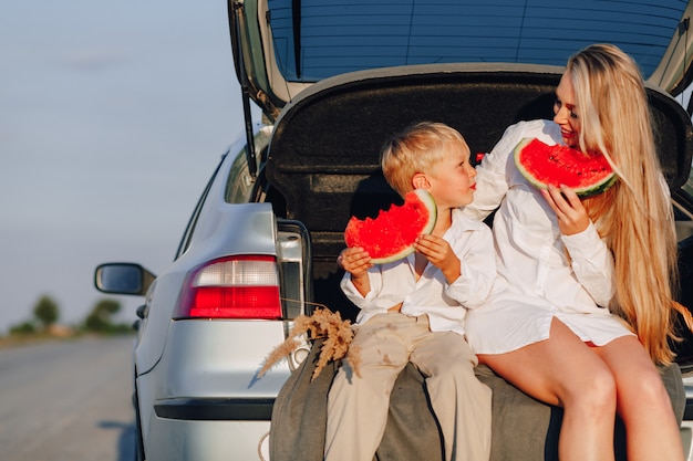 Hübsche blonde Haarfrau mit dem kleinen blonden Sohn bei Sonnenuntergang, der sich hinter dem Auto entspannt und Wassermelone isst. Sommer, Reisen, Natur und frische Luft auf dem Land.