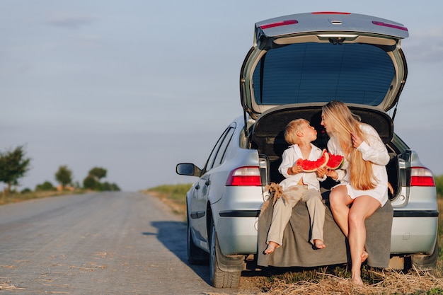 Hübsche blonde Haarfrau mit dem kleinen blonden Sohn bei Sonnenuntergang, der sich hinter dem Auto entspannt und Wassermelone isst. Sommer, Reisen, Natur und frische Luft auf dem Land.