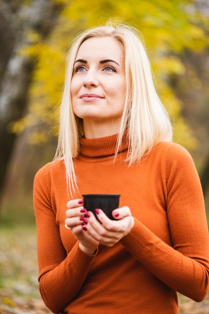 Hübsche blonde junge Frau mit blauen Augen in orangefarbenem Pullover hält schwarze Thermoskanne mit wärmendem Getränk auf gelbem Blätterhintergrund, helle Herbstsaison