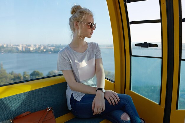 Hübsche blonde junge Frau mit Sonnenbrille, die auf dem Karussell sitzt und zur Seite schaut