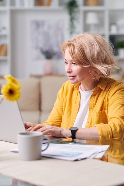 Hübsche blonde reife Frau in der gelben Jacke, die vom Schreibtisch vor Laptop während der Vernetzung in der häuslichen Umgebung sitzt