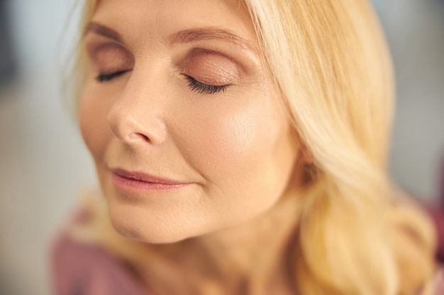 Foto hübsche blonde weibliche person, die ihre augen geschlossen hält, während sie lächelt und tief in ihren gedanken ist