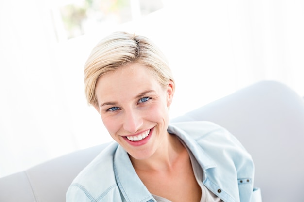 Hübsche Blondine, die auf der Couch sitzen und an der Kamera lächeln