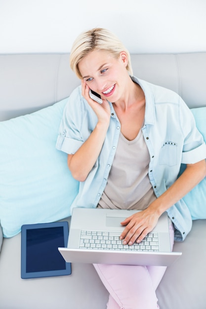 Hübsche Blondine, die ihren Laptop beim Ersuchen um das Telefon verwenden