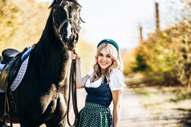 Hübsche Blondine in traditioneller Kleidung, die mit großem schwarzen Pferd draußen geht