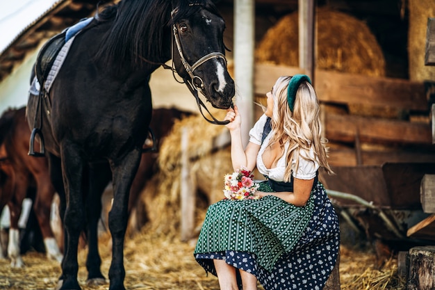 Hübsche Blondine in traditioneller Kleidung kümmert sich auf der Farm um das große schwarze Pferd