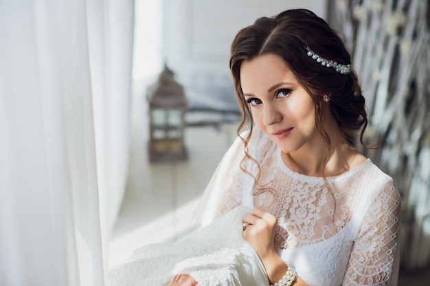 Hübsche brünette Braut im Seidenmantel und im Spitzenschleier, die Hochzeitskleid auf einem Kleiderbügel nahe dem Fenster halten.