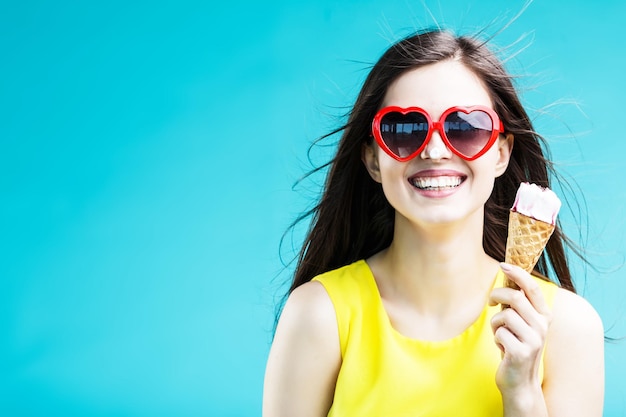 Hübsche brünette Frau in gelbem Hemd und Sonnenbrille, die vor blauem Hintergrund Waffeleis isst