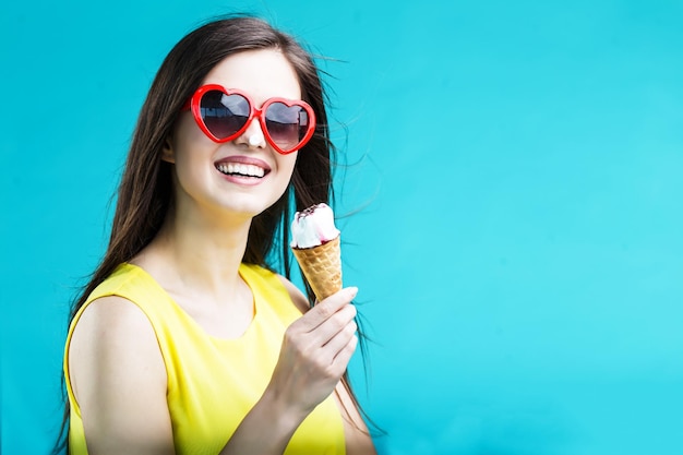 Hübsche brünette Frau in gelbem Hemd und Sonnenbrille, die vor blauem Hintergrund Waffeleis isst