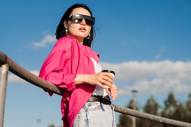 Hübsche brünette Frau mit Sonnenbrille und Freizeitkleidung, die eine Kaffeetasse hält und über dem blauen Himmel im Park posiert