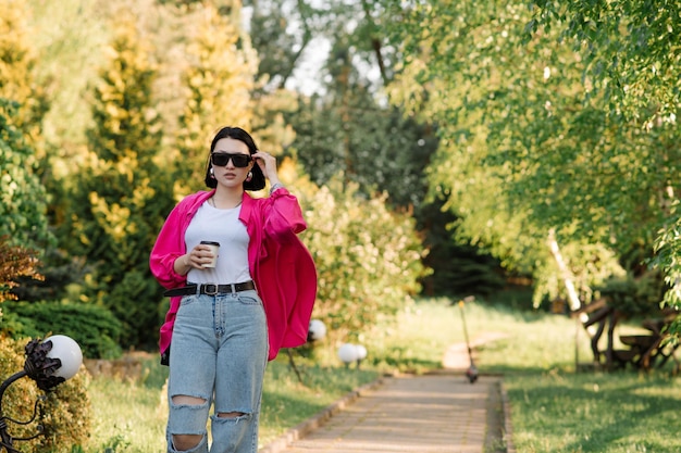 Hübsche brünette Frau mit Sonnenbrille und Kaffeetasse, die im Park spazieren geht
