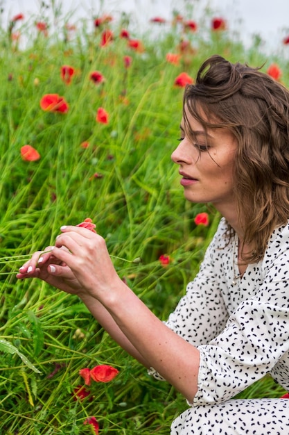 Hübsche Brünette mit lockigem Haar, die ein elegantes Kleid trägt, spielt mit roter Mohnblume in der Hand