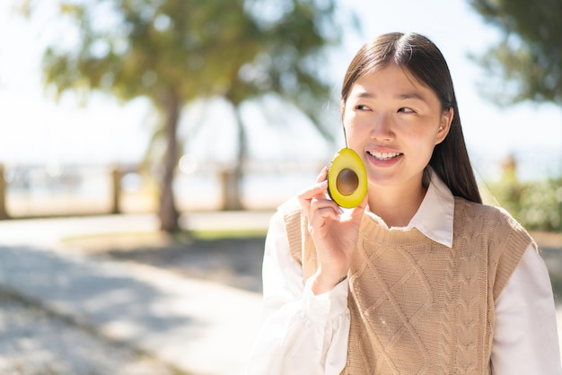 Hübsche Chinesin im Freien, die eine Avocado mit glücklichem Ausdruck hält