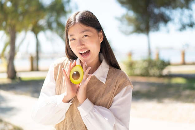 Hübsche Chinesin im Freien, die eine Avocado mit überraschtem Ausdruck hält