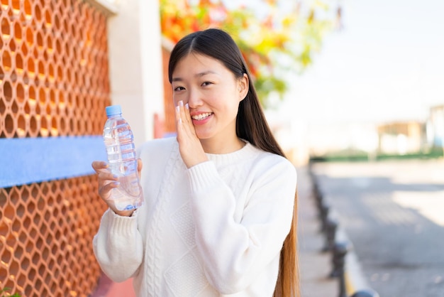 Hübsche Chinesin mit einer Flasche Wasser im Freien, die etwas flüstert