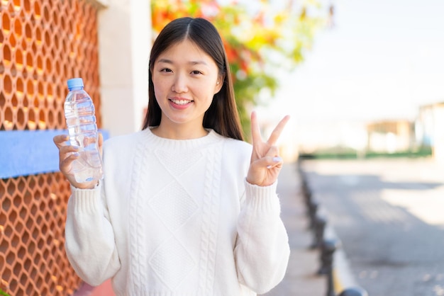 Hübsche Chinesin mit einer Flasche Wasser im Freien, lächelnd und mit Siegeszeichen